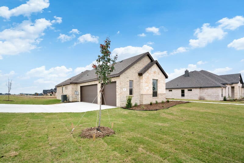 New single-story home with light brick exterior, two-car garage, and freshly landscaped yard in a suburban neighborhood under a sunny sky.
