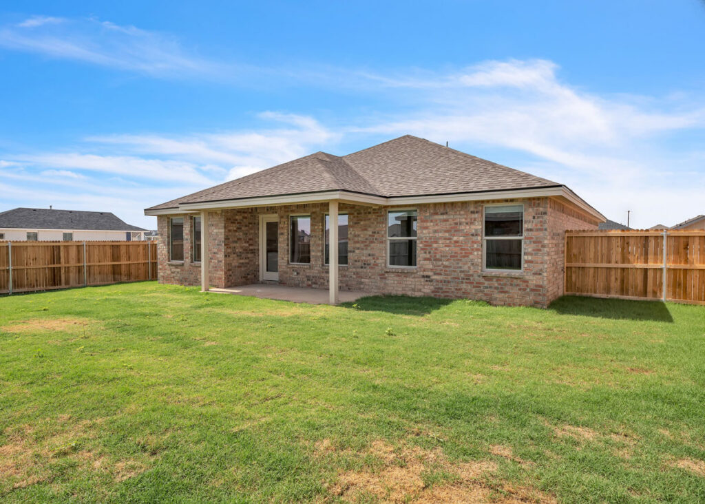 Back exterior of a single-story brick home by Silver Leaf Homes, featuring a covered patio, large fenced backyard with green grass, and neutral shingle roof under a clear blue sky.